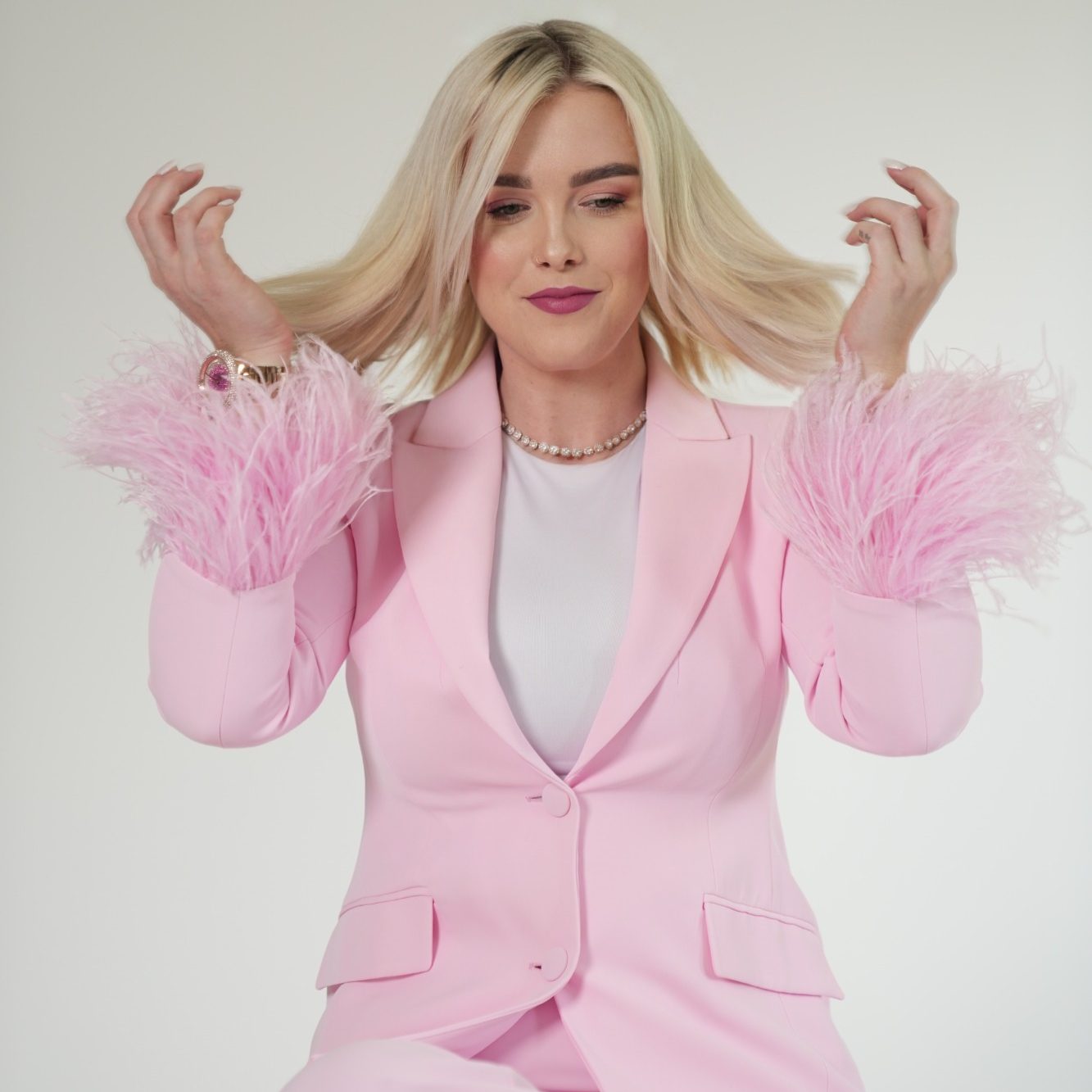 Jenny Martens – Mobile Friseurmeisterin in München Jenny Martens, mobile Friseurin in München, in einem pinken Anzug mit auffälligen, gefiederten Ärmeln und langen blonden Haaren.